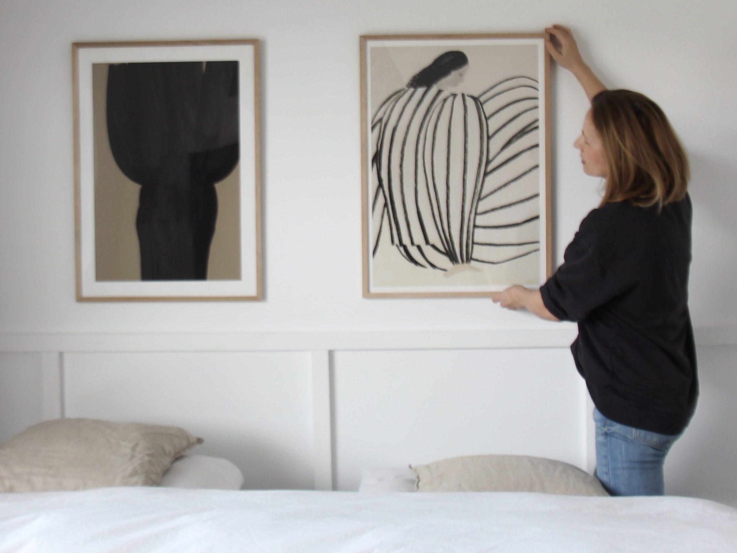 Eine Person hängt ein Kunstwerk an eine Wand in einem hellen Raum mit einem Bett im Vordergrund und mehreren Kissen.